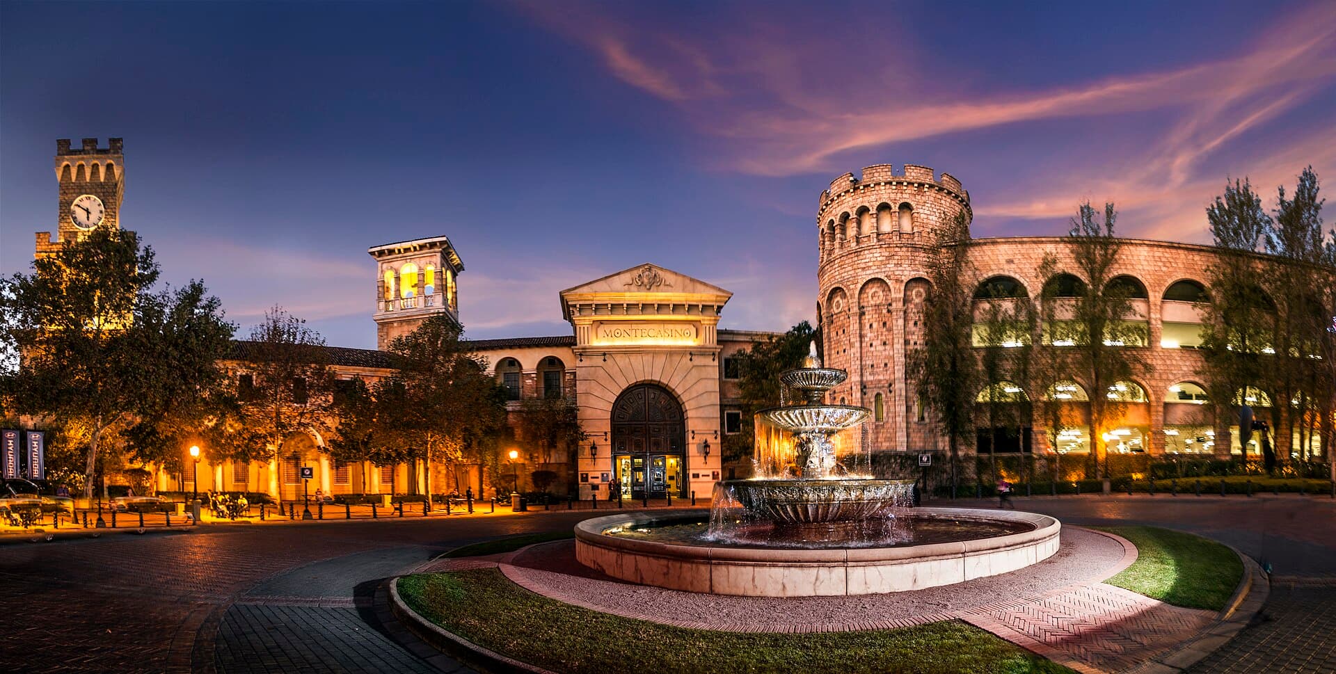 Montecasino's mock-Tuscan facades, clock tower and stone rotunda glowing at dusk behind a tiered fountain in the entrance piazza.